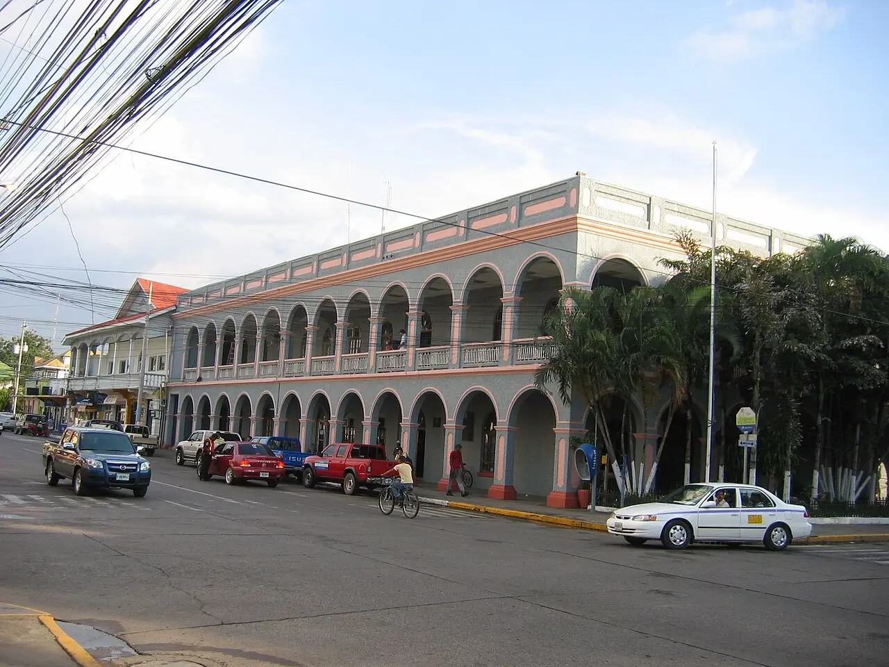 Edificio de dos pisos con arcos en la planta baja y en el balcón del segundo piso, pintado de gris con detalles rosados, palmeras a la derecha, carros y un taxi blanco en la calle y una persona en bicicleta