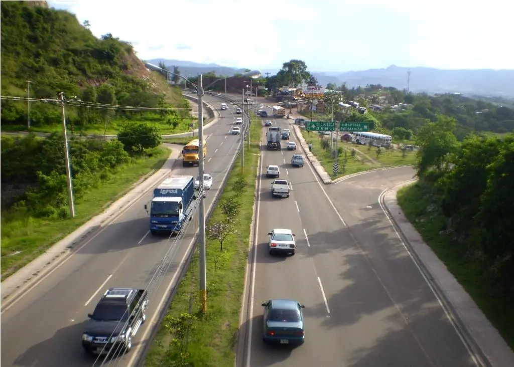Carros, un camión y un bus escolar circulando por una carretera de cuatro carriles entre cerros verdes