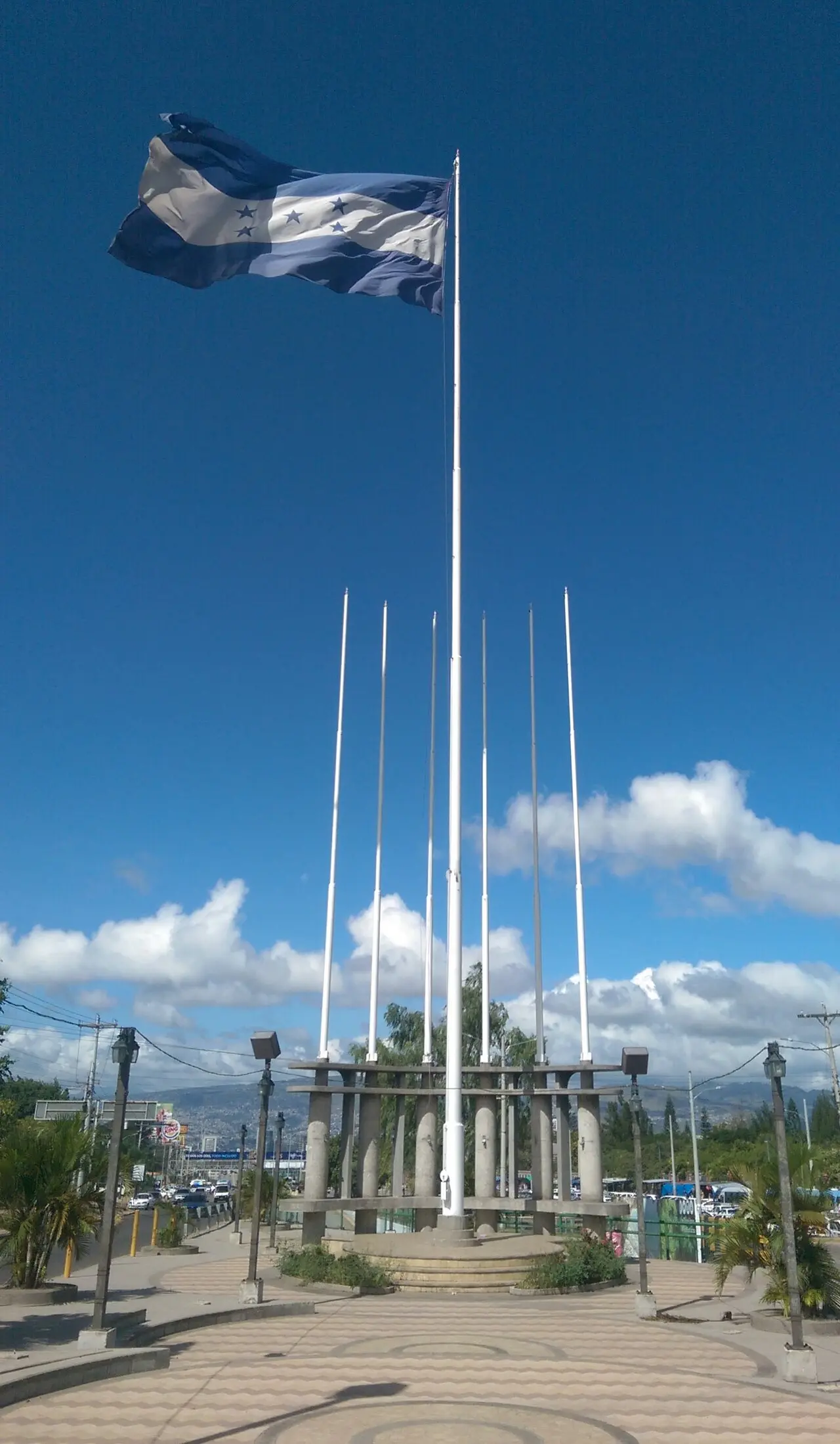 Bandera de Honduras de franjas azul oscuro y cinco estrellas ondeando en lo alto de un asta blanca, sobre una plaza con seis astas vacías, faroles y cielo azul con nubes