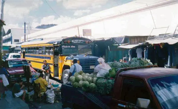 Calle de mercado en Comayagüela con puestos de verduras, un picop cargado de sandías y repollos y un bus amarillo