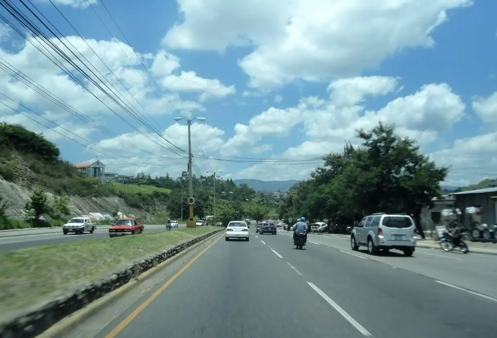 Carros y una moto circulando por un bulevar de cuatro carriles en Tegucigalpa, con cerros al fondo