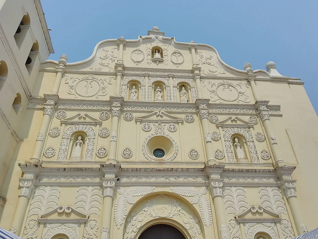 Fachada barroca de color crema de la catedral de Comayagua, con columnas, relieves de hojas y rosetas, santos de piedra en nichos y una ventana redonda sobre la puerta, bajo un cielo azul