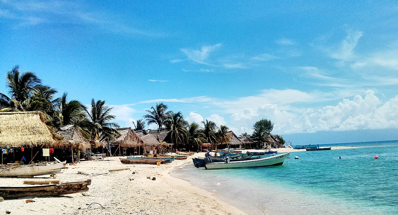 Playa de arena blanca con palmeras, ranchos de techo de manaca y lanchas de pescadores varadas y flotando en agua turquesa, en el cayo Chachahuate