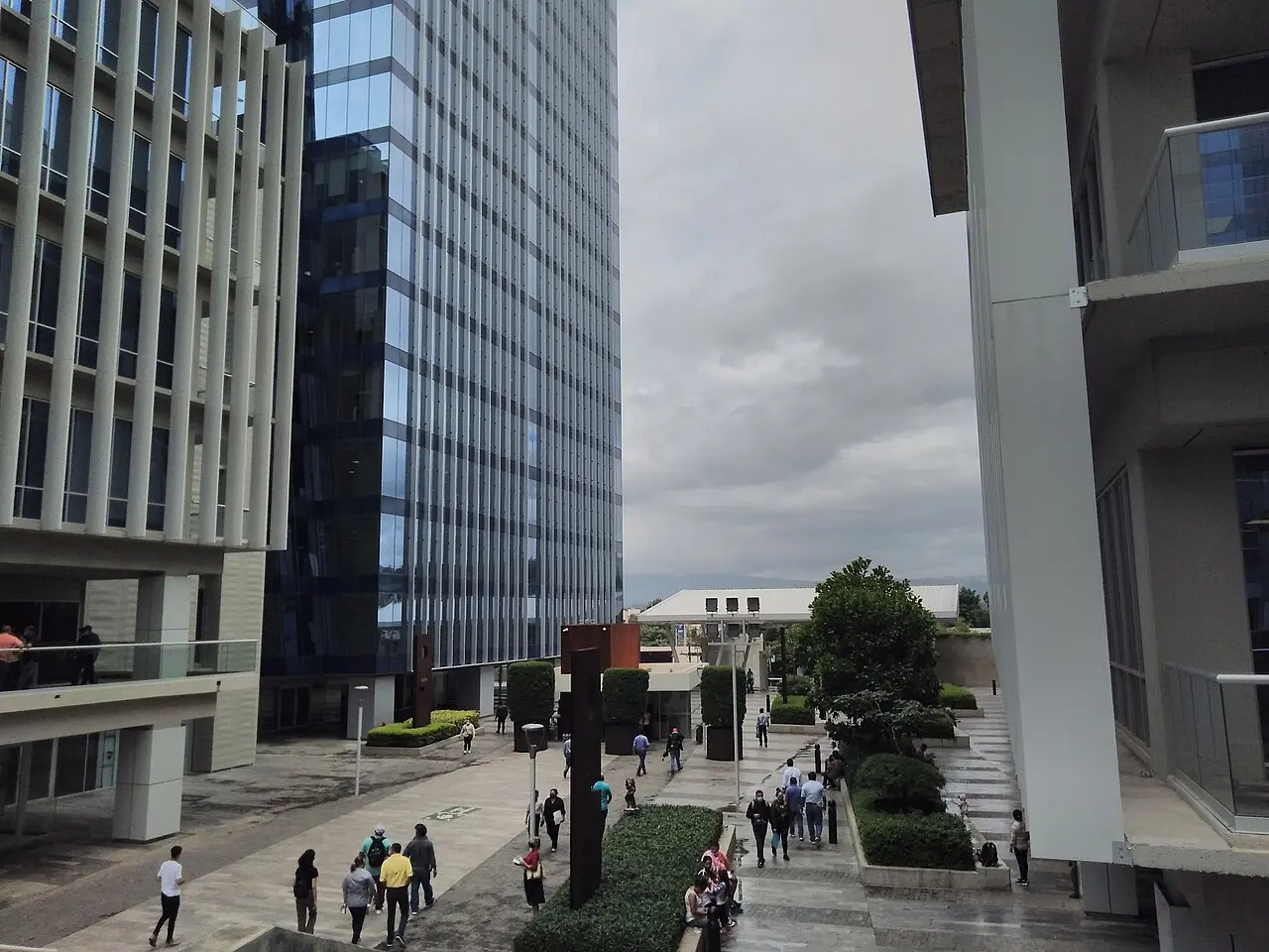 Edificios de vidrio y concreto del Centro Cívico Gubernamental en Tegucigalpa, con un paseo peatonal con jardineras y gente caminando, bajo un cielo nublado