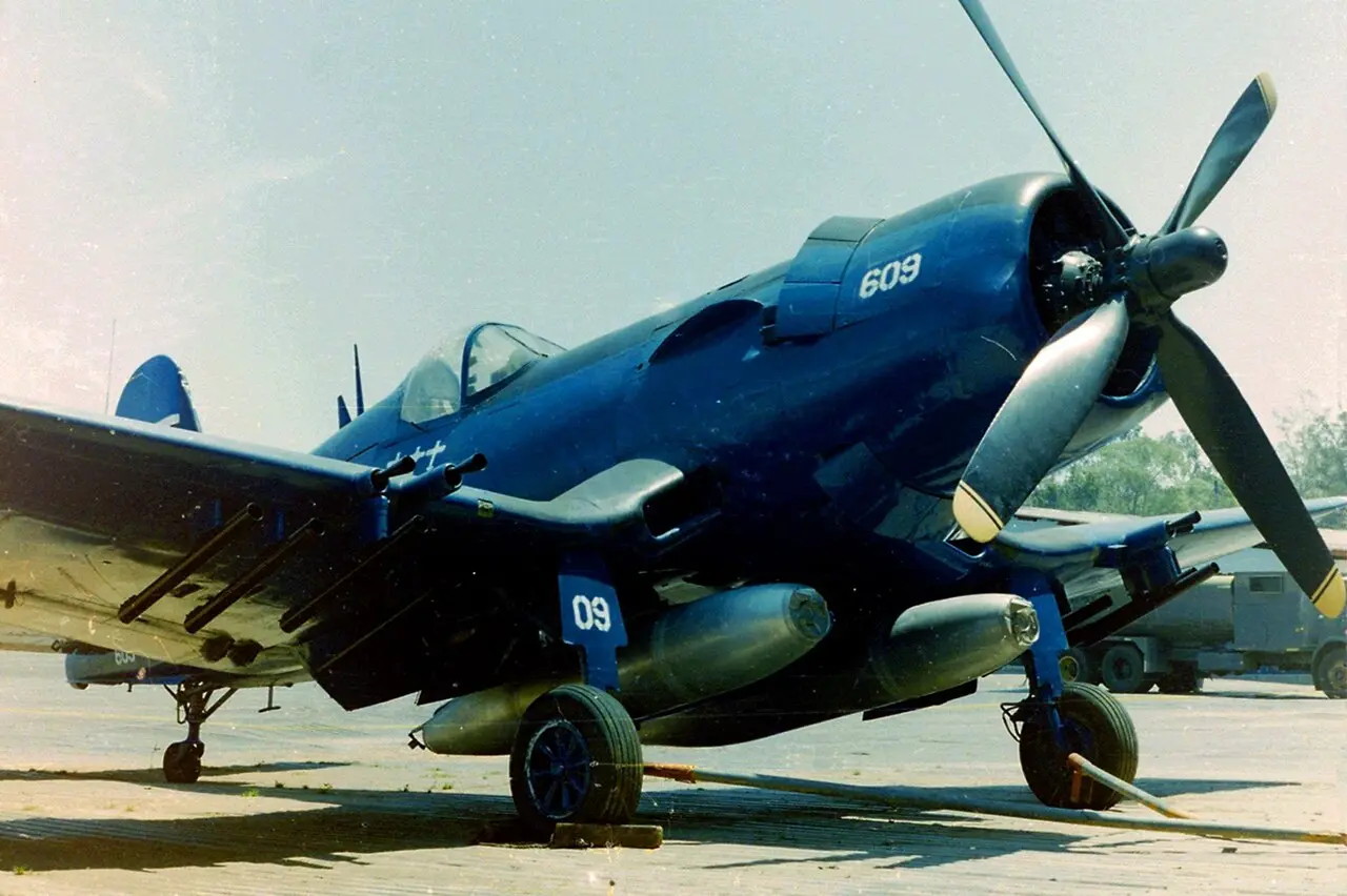 Avión militar de hélice pintado de azul oscuro, con el número 609 en el motor, cuatro palas en la hélice, cañones en las alas y tanques bajo el fuselaje, estacionado sobre una pista de concreto con árboles y un camión al fondo