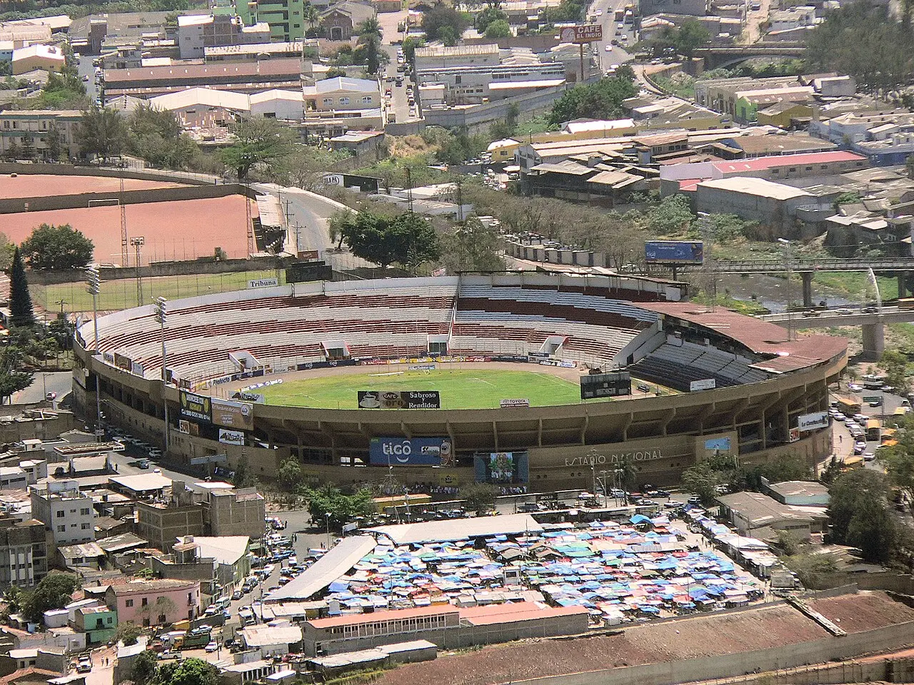 Estadio de fútbol ovalado visto desde un cerro, con graderías blancas y rojas, cancha verde y el letrero Estadio Nacional en la fachada; alrededor hay casas, calles y un mercado con toldos azules
