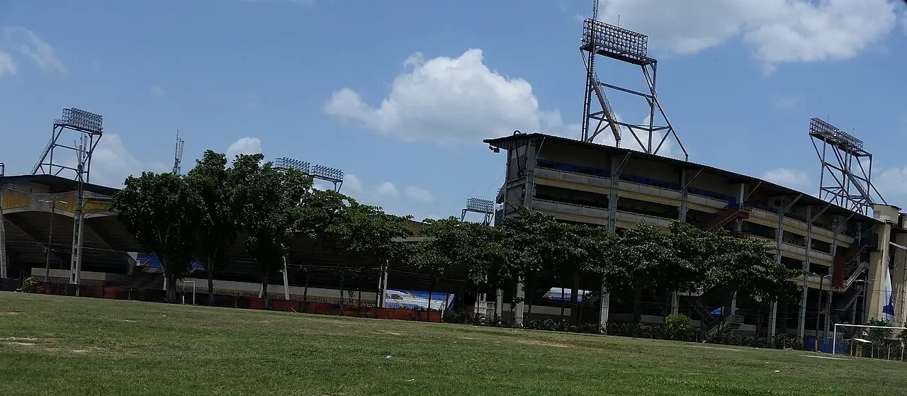 Exterior de un estadio de concreto con graderías techadas y torres de iluminación altas, detrás de una hilera de árboles y un campo de grama, bajo un cielo azul con nubes