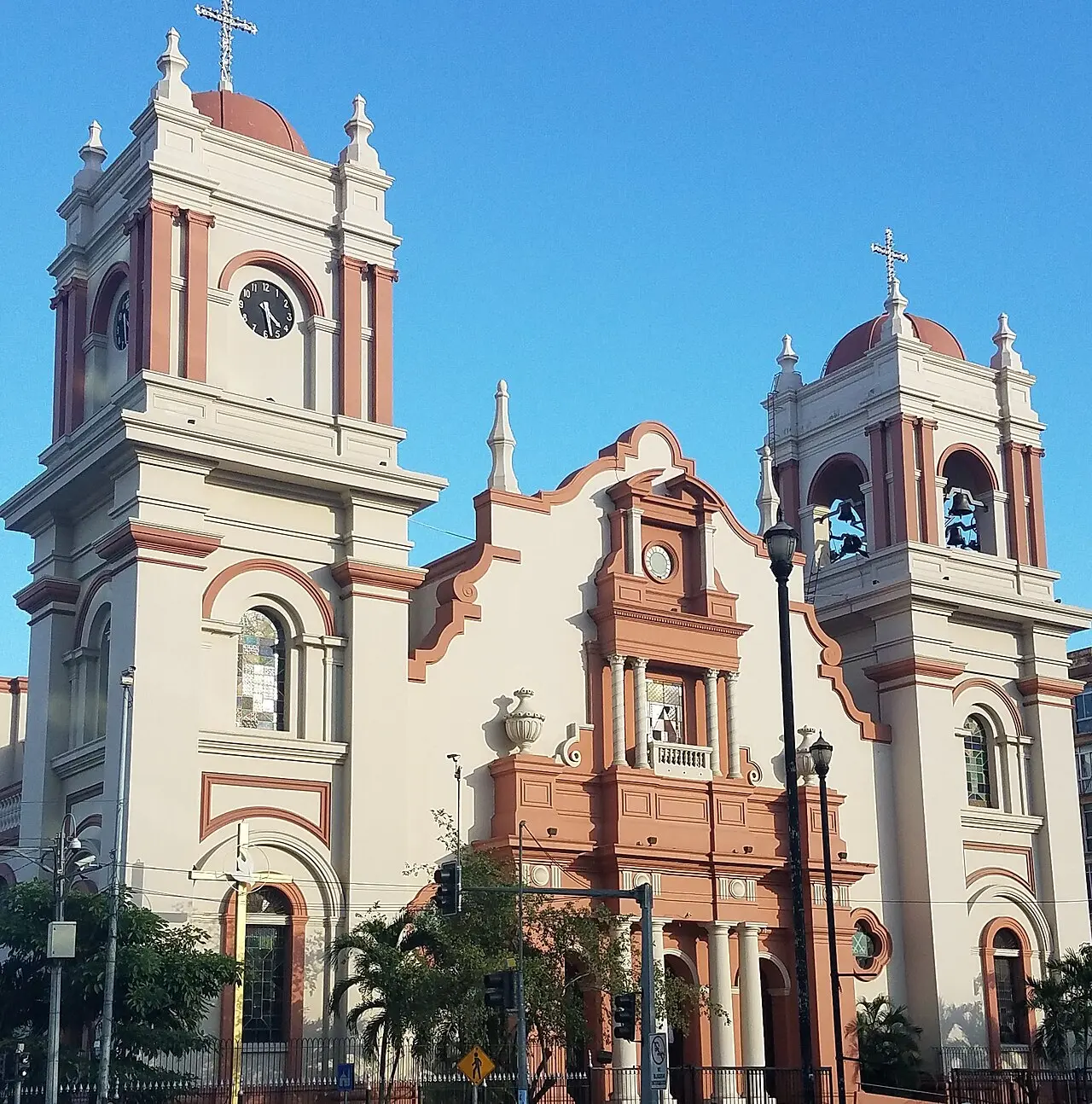 Fachada de la Catedral de San Pedro Apóstol, de color crema con detalles terracota, dos torres con cúpulas y cruces, un reloj en la torre izquierda y campanas en la derecha, bajo un cielo azul