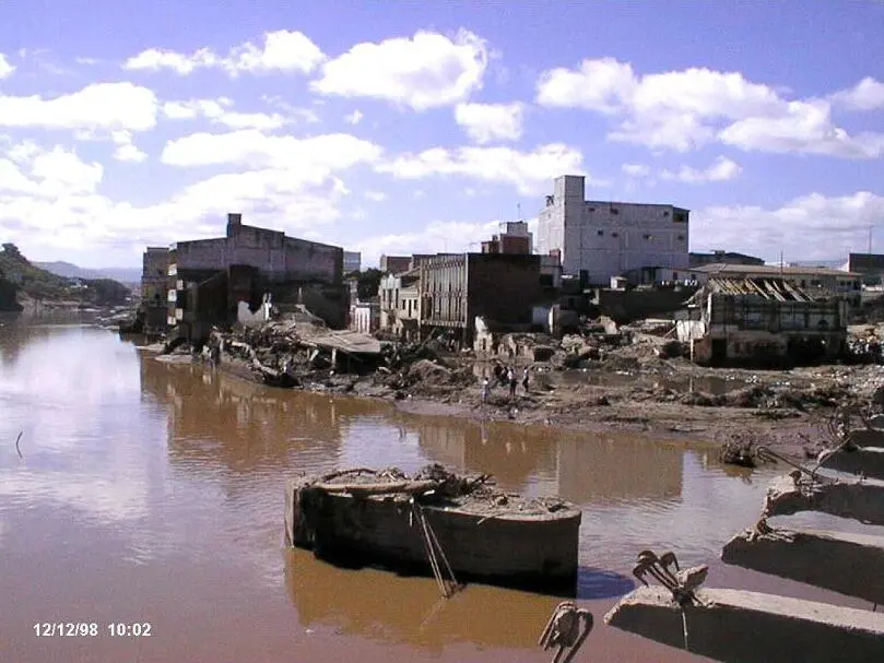 Río café y crecido junto a edificios dañados y escombros a la orilla, con restos de un puente en primer plano y algunas personas caminando entre el lodo