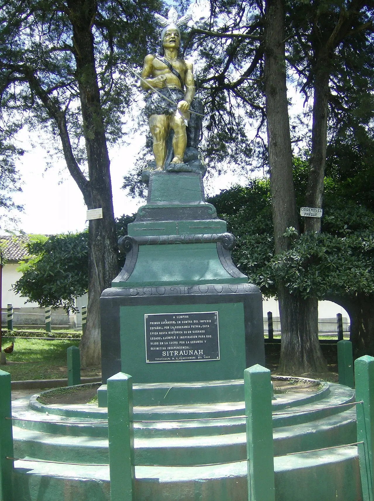Estatua dorada de un guerrero indígena con penacho, arco y flecha, sobre un pedestal verde y negro con una placa, rodeada de postes verdes y árboles altos