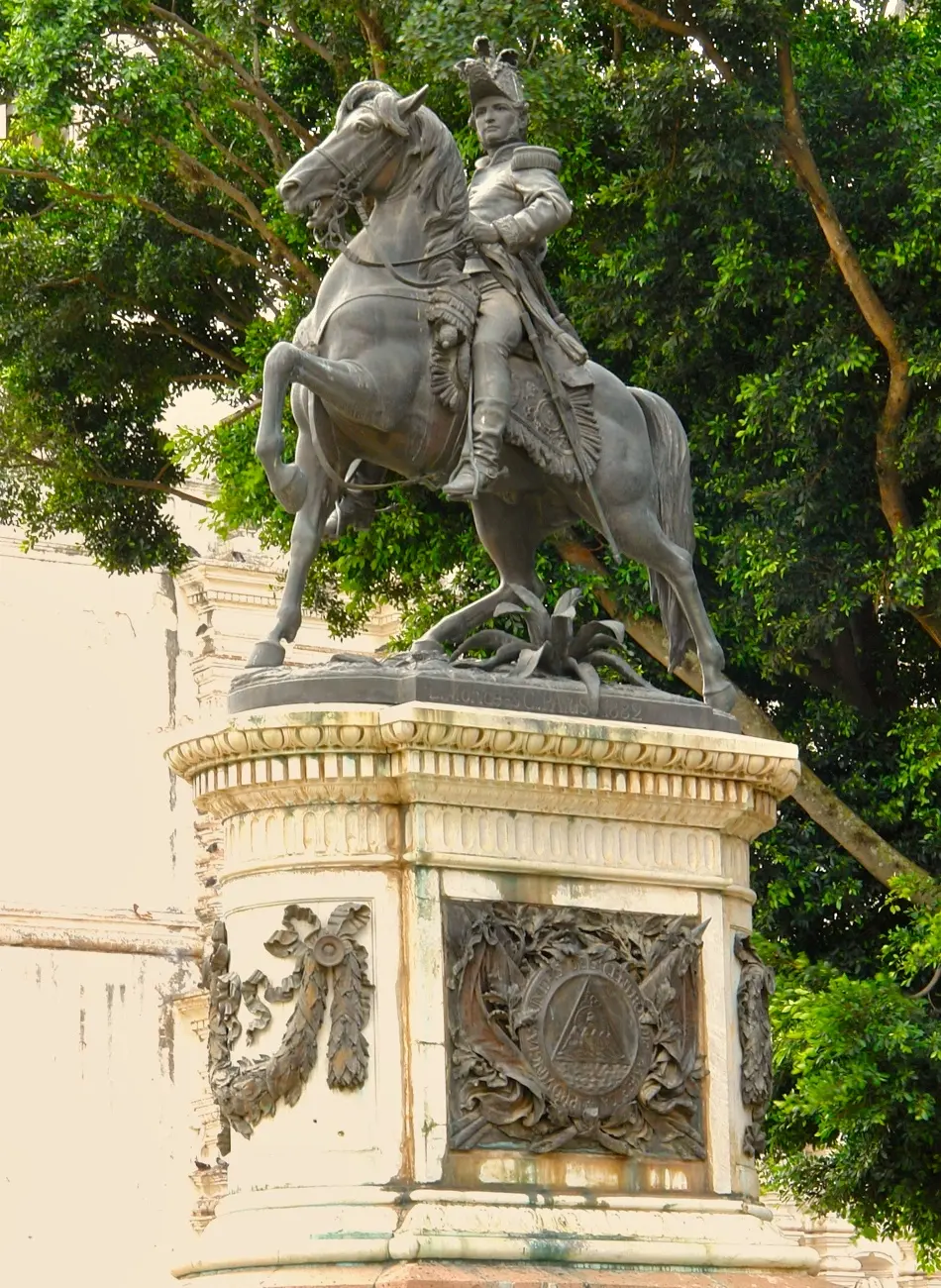 Estatua ecuestre de bronce de Francisco Morazán con sombrero militar sobre un caballo encabritado, en lo alto de un pedestal blanco con un escudo en relieve, frente a árboles frondosos