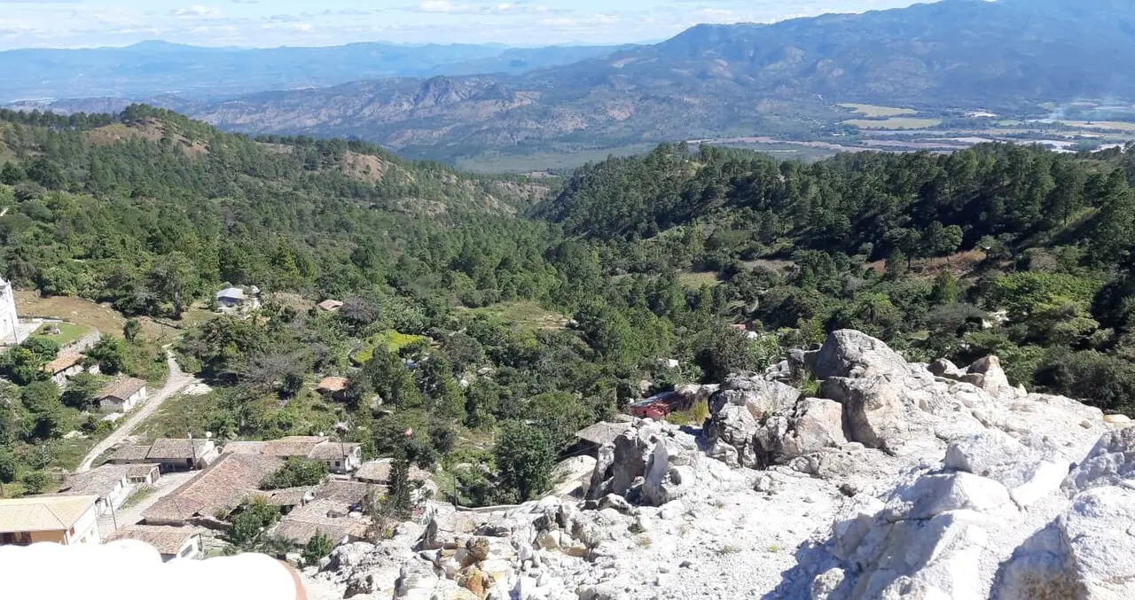 Vista desde lo alto de un pueblo de casas con techos de teja entre pinares, con rocas blancas en primer plano y montañas y un valle al fondo