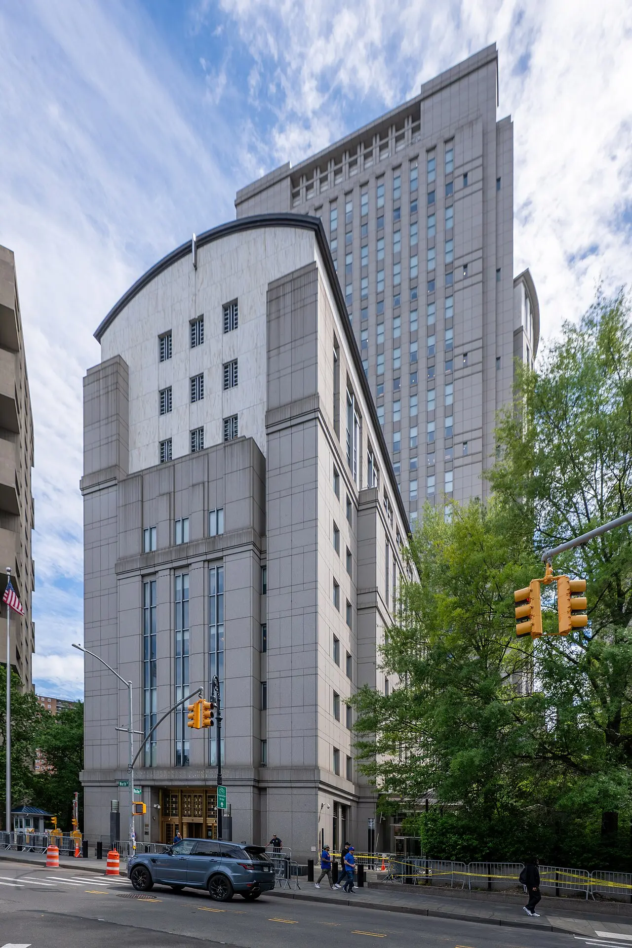 Edificio alto de granito gris con entrada de puertas doradas, una torre detrás, árboles a la derecha, semáforos amarillos y una camioneta en la calle frente al edificio