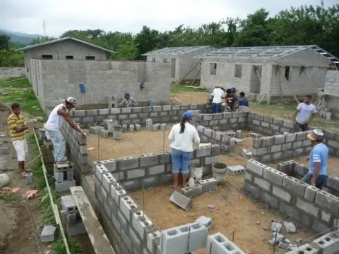 Obra en construcción con paredes de bloque gris a medio levantar, varios trabajadores colocando bloques y, al fondo, casas de bloque ya techadas entre árboles